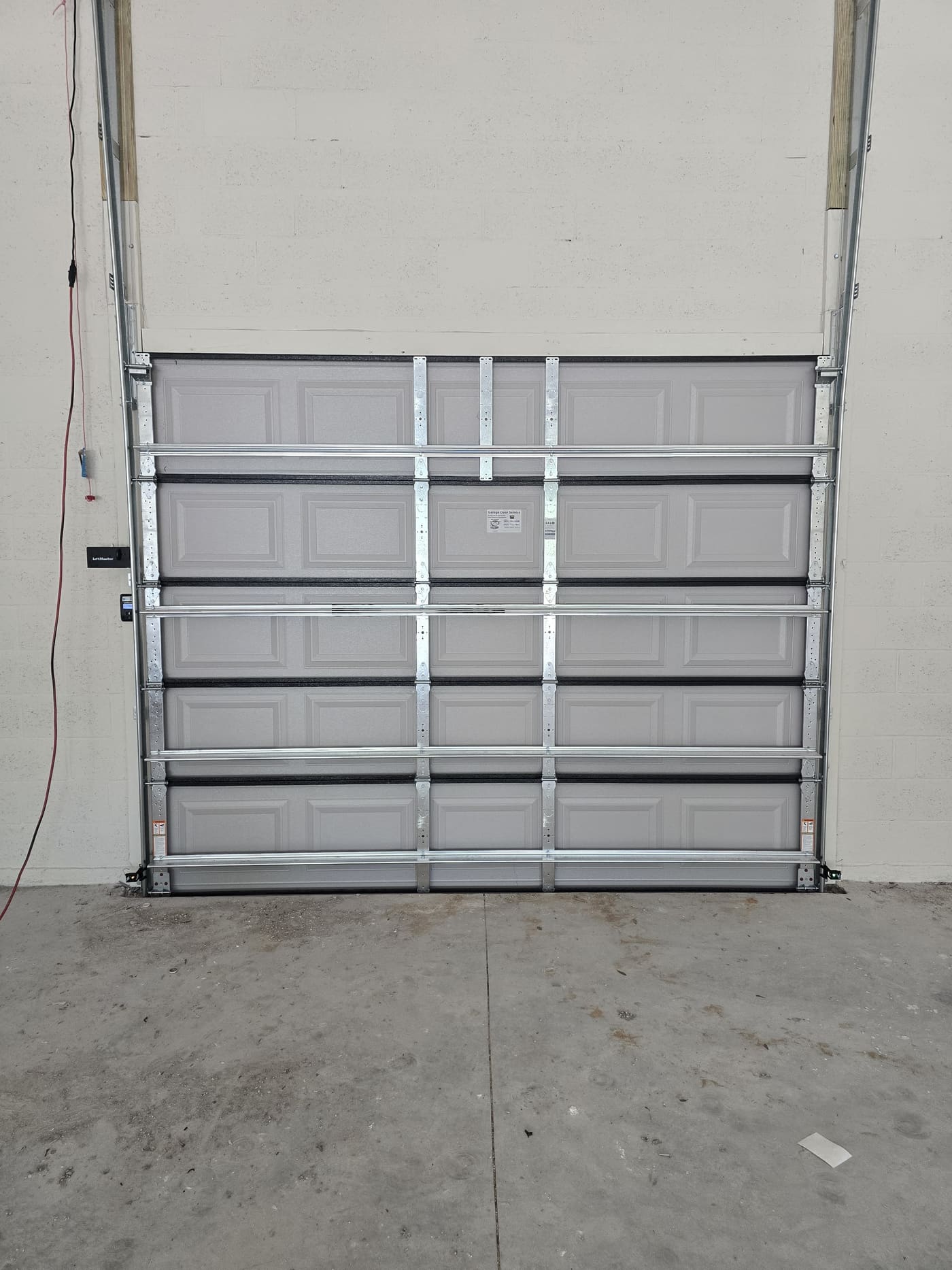 A grey raised-panel garage door seen from inside a block-walled garage, with steel struts across it, the vertical tracks either side and a wall control on the left.